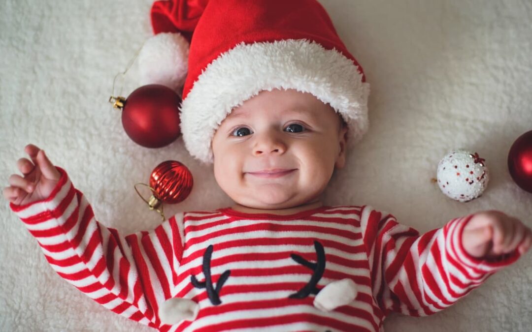 Bebé con gorro de Navidad y jersey de rayas rojas, rodeado de adornos festivos.