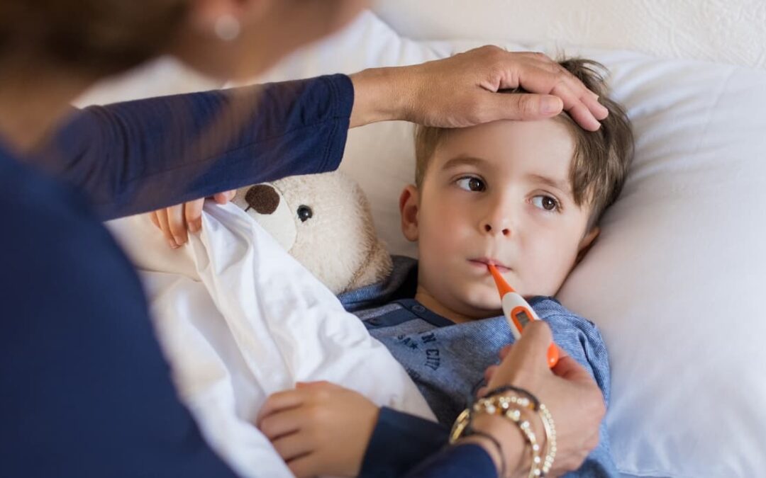 Niño acostado en cama con termómetro en la boca, acompañado de su peluche, mientras una mujer verifica su temperatura tocándole la frente.