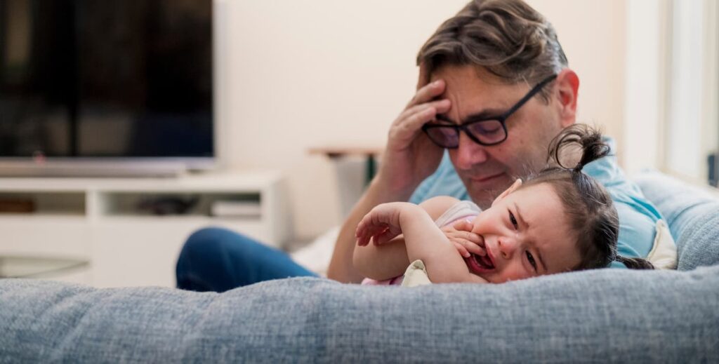 Un padre muestra paciencia mientras su hijo, visiblemente frustrado, tiene un berrinche en el suelo. El padre, con una expresión serena, está sentado a su lado, intentando consolarlo y entender sus sentimientos en un ambiente hogareño.